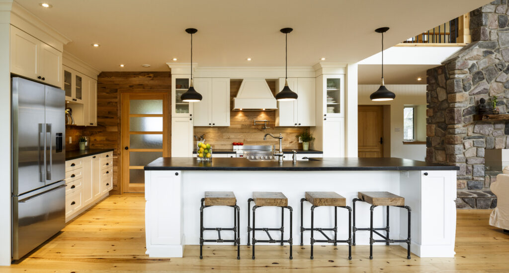 Modern kitchen with white cabinets, stainless steel appliances, a large island with four wooden bar stools, black pendant lights, wooden floors, and a stone accent wall.