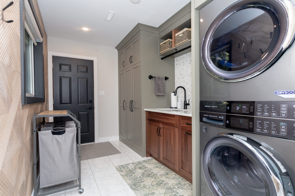 Modern laundry room with a stacked washer and dryer, wood and green cabinetry, a black faucet, a patterned rug, a laundry hamper, and a black door at the back. The space is tidy and well-organized.