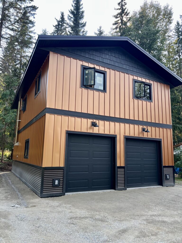 A modern two-story building with copper-colored vertical siding and two black garage doors, surrounded by tall evergreen trees. The structure has black trim and windows on the upper level.