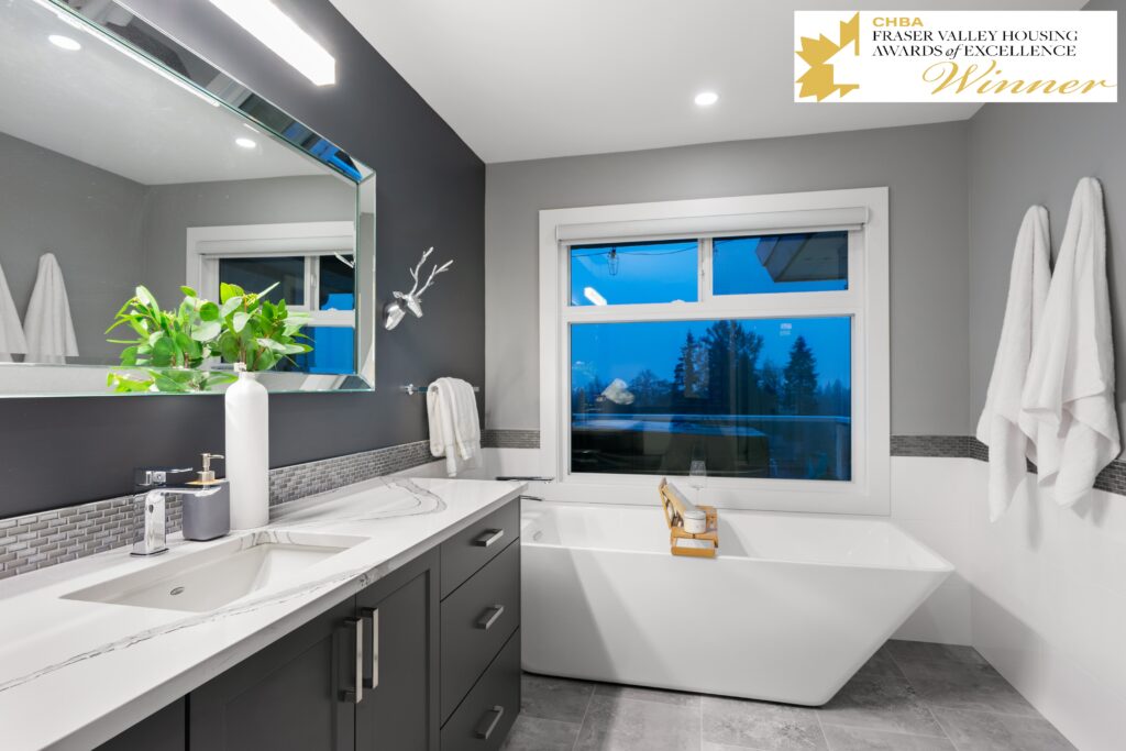 Modern bathroom with gray cabinets, white countertop, large mirror, and decorative plant. A white freestanding bathtub sits by a window with blue evening light. Award badge is displayed in the top right corner.