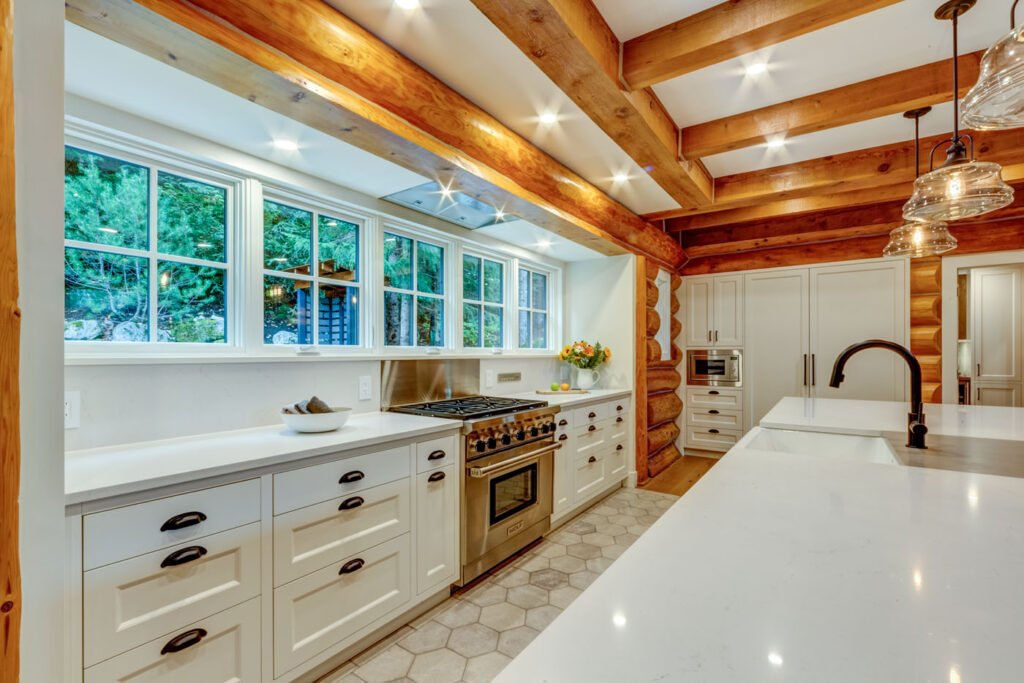 A spacious, modern log cabin kitchen with exposed wood beams, white cabinetry, stainless steel appliances, and large windows overlooking greenery. Quartz countertops and pendant lights complete the bright, airy space.