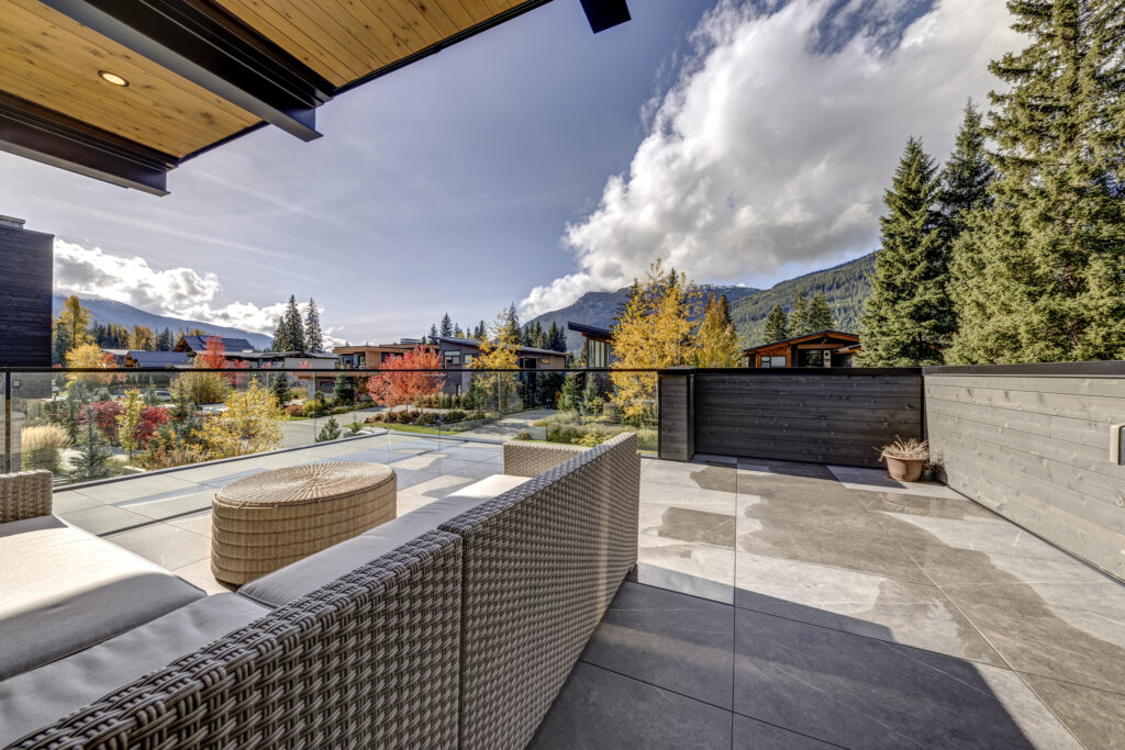A spacious modern terrace with a wicker sofa and round rattan table overlooks a landscaped garden, mountain views, and autumn trees under a partly cloudy sky. Glass railing lines the terrace.