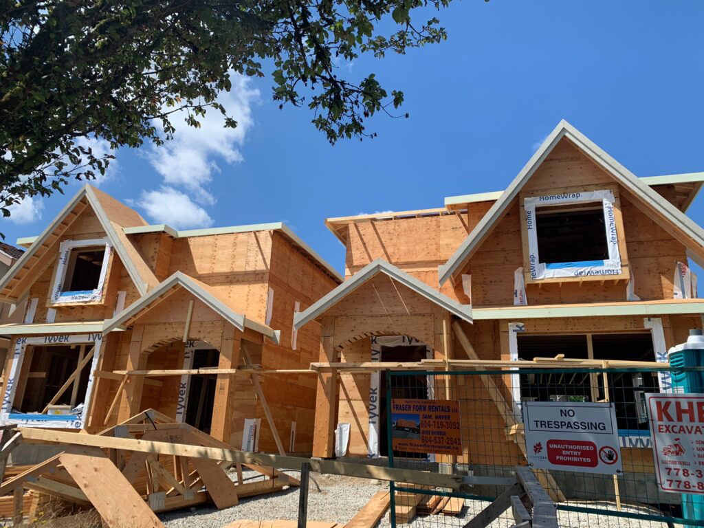 Two partially built wooden houses with peaked roofs under construction, surrounded by scaffolding and fencing. Signs on the fence include No Trespassing and construction company information. A tree branch is visible overhead.