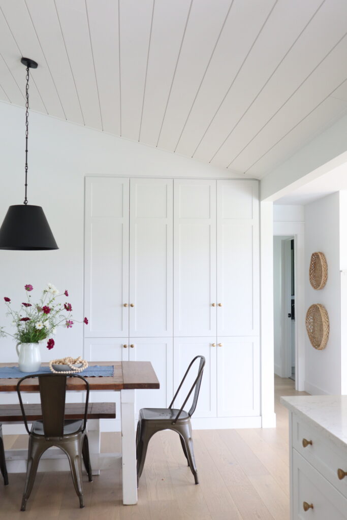 A bright, modern kitchen with white paneled cabinets, light wood flooring, a wooden dining table with metal chairs, a black pendant light, a vase of flowers, and woven baskets hanging on the wall.