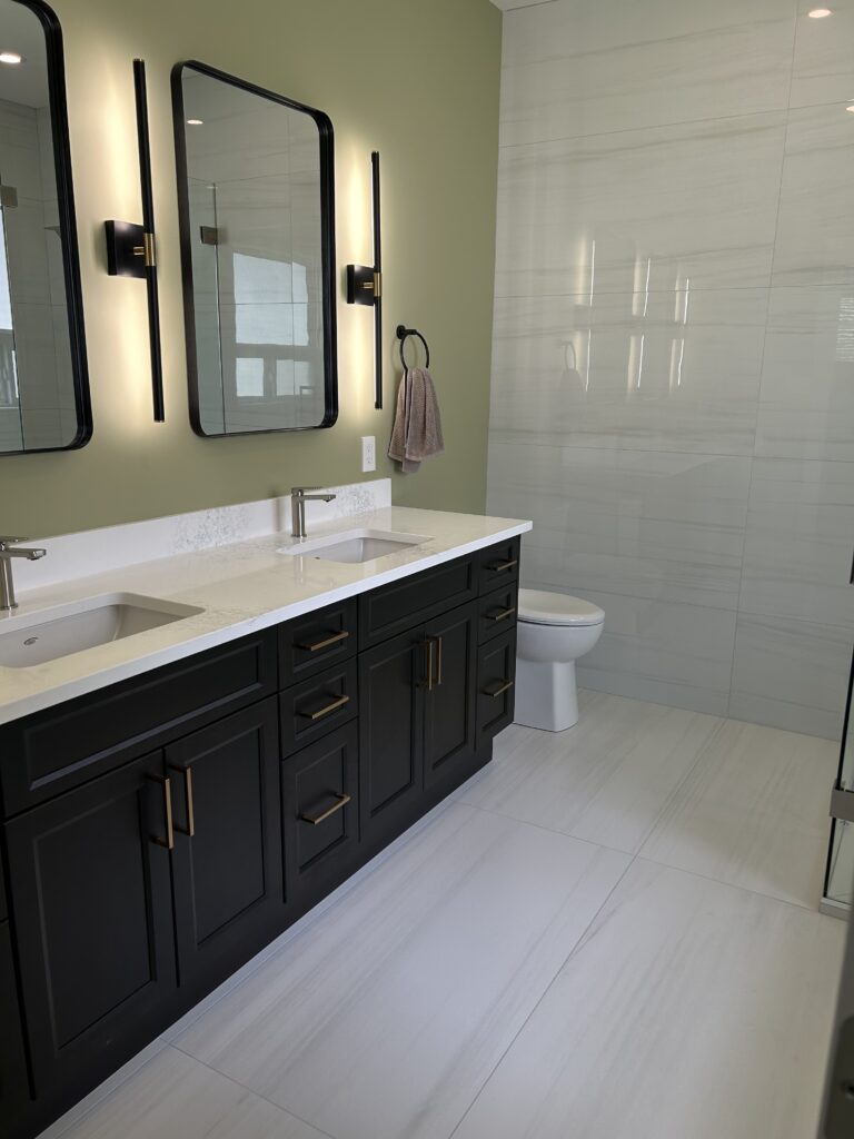 Modern bathroom with a double-sink vanity featuring dark cabinets, white countertop, rectangular mirrors with black frames, wall-mounted lights, green accent wall, towel ring, and a toilet against a tiled wall and floor.