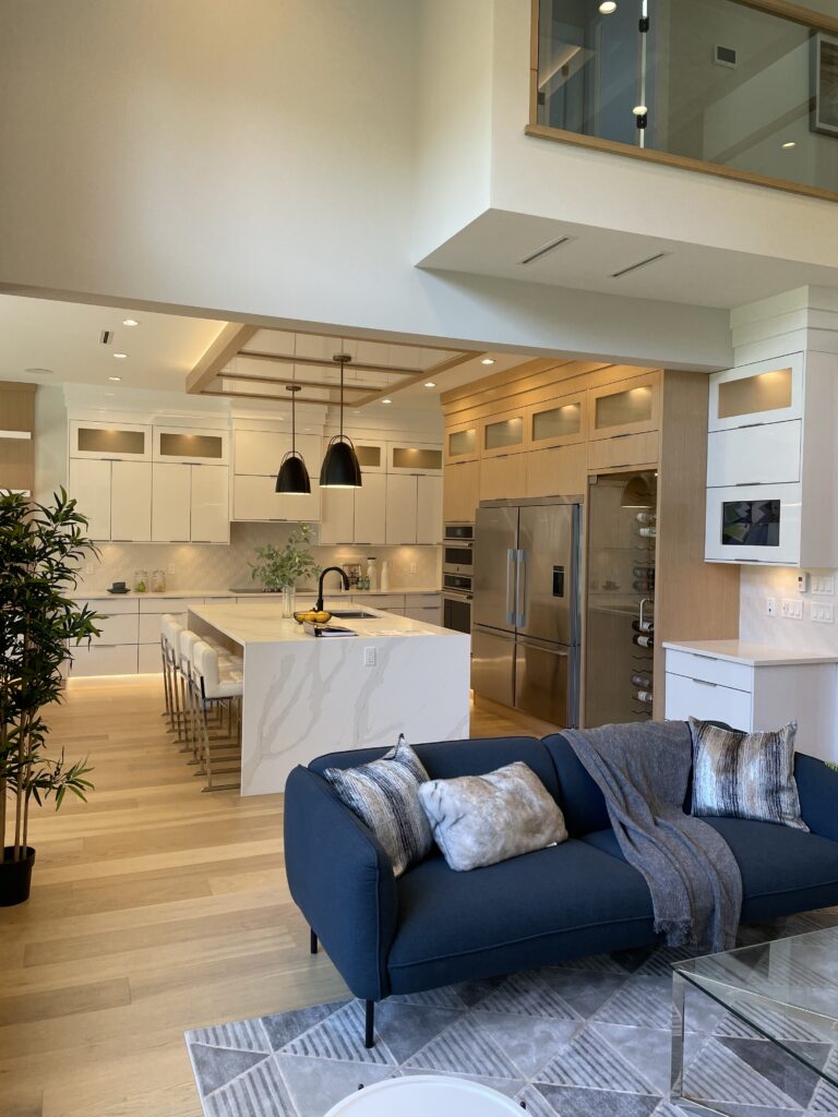 Modern open-concept kitchen with white cabinets, marble island, and pendant lights. A blue sofa with pillows and a throw blanket sits on a patterned rug in the living area. Light wood floors and large windows brighten the space.