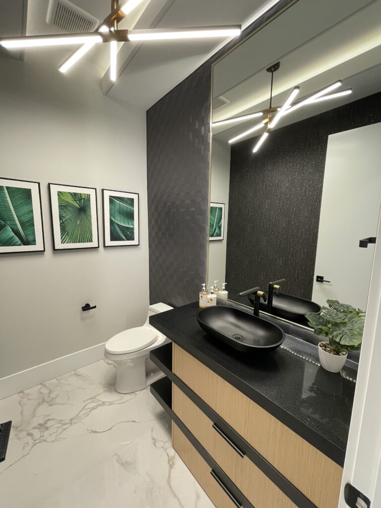 Modern bathroom with black fixtures, a black vessel sink, wood vanity, marble floor, patterned dark accent wall, and three framed leaf prints. A potted plant and soap bottles sit beside the sink under geometric lights.