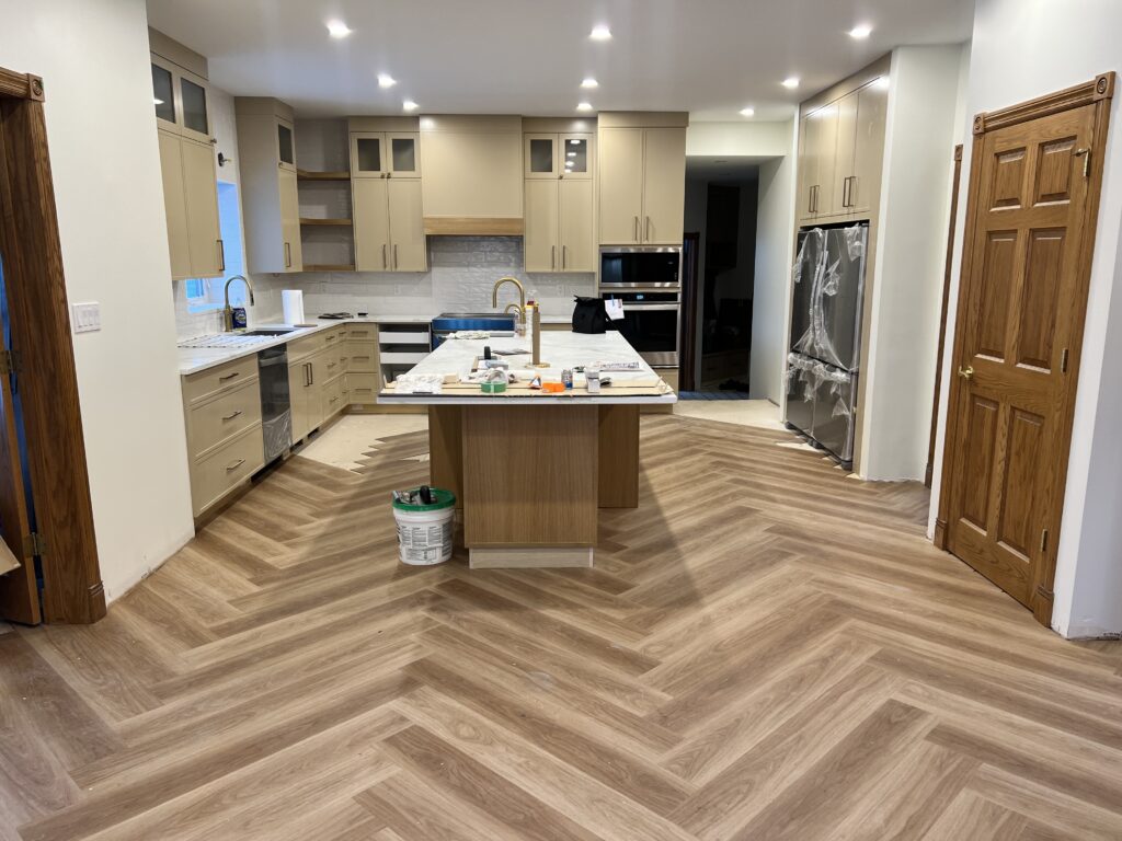 A modern kitchen under renovation with light wood cabinets, a central island, built-in ovens, and herringbone-patterned wood flooring. Tools and supplies are on the island, and appliances are partially covered in plastic.