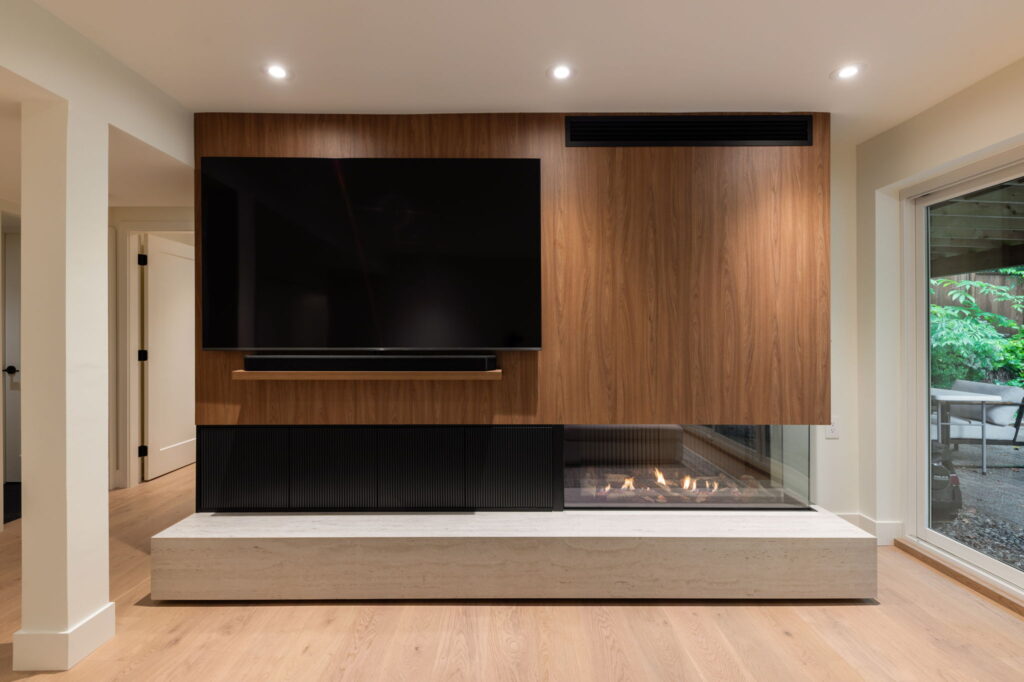 Modern living room with a large wall-mounted TV above a sleek black console, a horizontal gas fireplace, wood paneling, and a large window showing greenery outside. Light wood flooring and recessed ceiling lights complete the space.