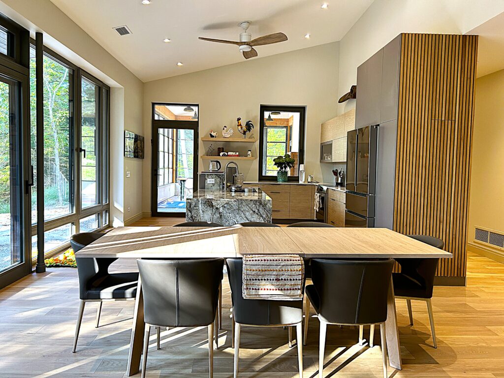 Modern kitchen and dining area with large windows, wooden floors, black chairs around a wooden table, and a marble kitchen island. Natural light fills the space, highlighting sleek cabinets and minimalist decor.