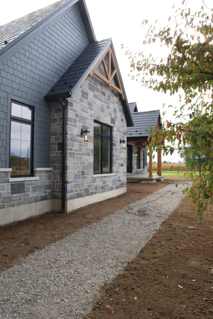 A modern house with gray stone walls, dark roof shingles, and large windows sits beside a gravel pathway; bare soil and a tree with green leaves are visible in the foreground.