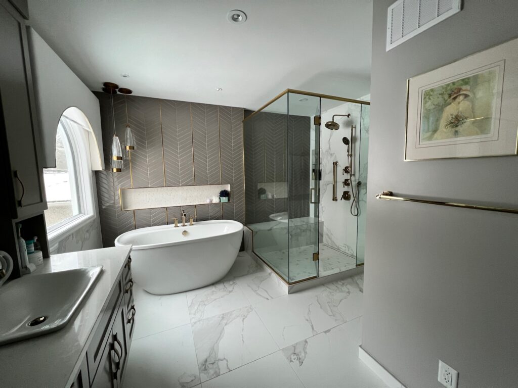 Modern bathroom with a freestanding white tub, glass-enclosed shower with gold fixtures, patterned tile accent wall, marble-style floor, wall-mounted sink, and framed artwork on a gray wall. Natural light streams in from a window.