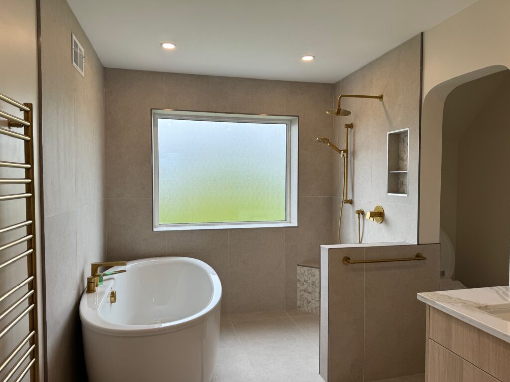 Modern bathroom with a freestanding tub, a large frosted window, a walk-in shower with gold fixtures, a built-in shower bench, a heated towel rack, and light beige tiles throughout.