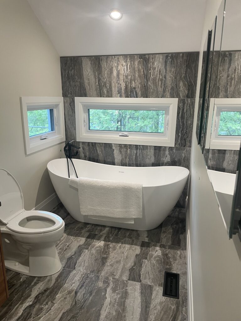 Modern bathroom with a freestanding white bathtub, towel draped over the side, nearby toilet, marble-patterned tiles on the wall and floor, and two rectangular windows letting in natural light.