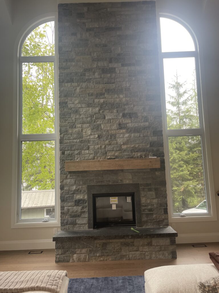 A tall stone fireplace with a wooden mantel is centered between two large arched windows, letting in natural light and showing green trees outside. The room has light walls, a blue rug, and beige furniture.