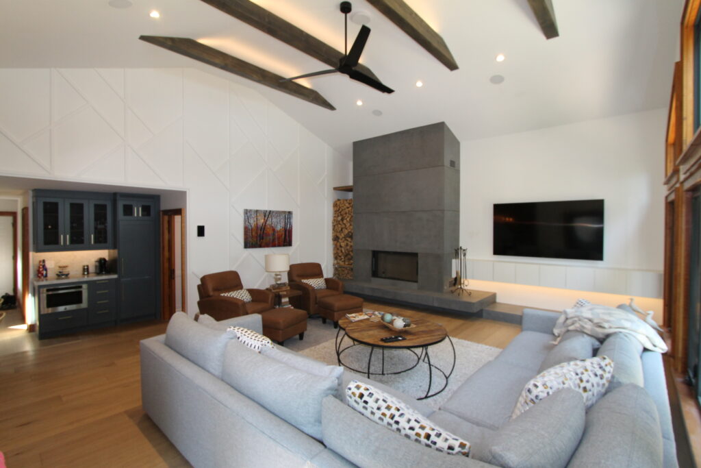 A modern living room with a large gray sectional sofa, round coffee table, two brown armchairs, a minimalist fireplace with concrete surround, built-in shelves, and a flat-screen TV on a white wall. Exposed wooden beams and ceiling fan above.