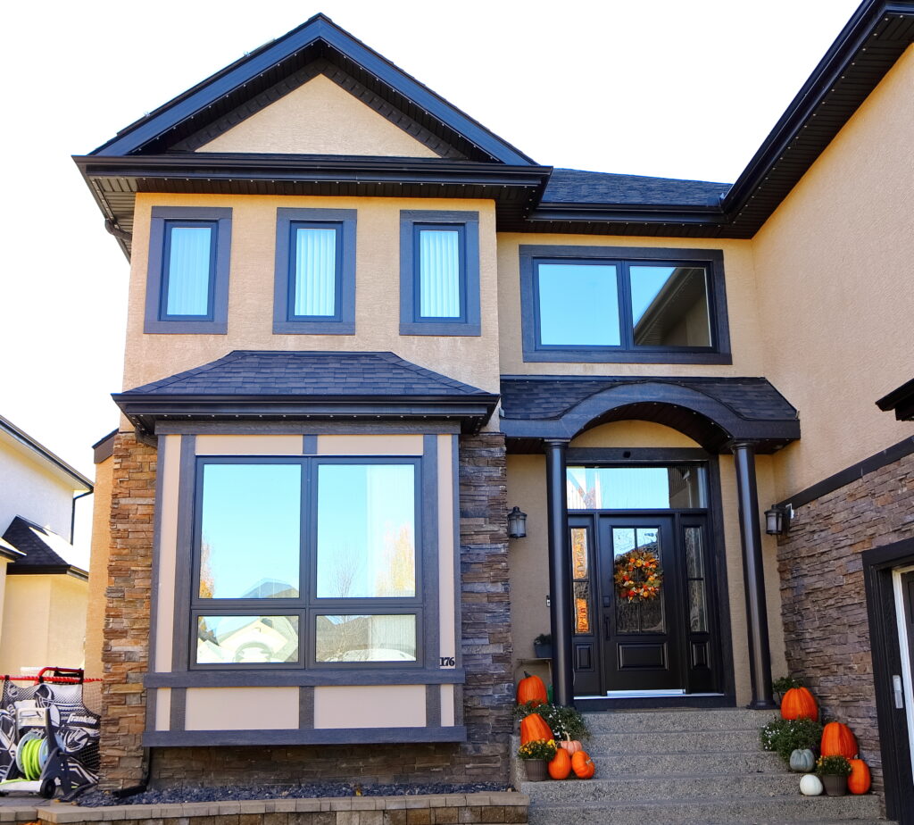 Two-story house with beige walls, dark trim, and stone accents. The front porch is decorated with pumpkins and a fall wreath on the door, suggesting autumn or Halloween season.
