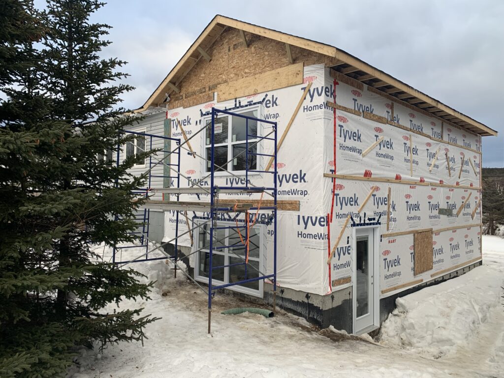 A house under construction in a snowy area, with Tyvek wrap covering the exterior walls, scaffolding set up by large windows, and unfinished roof edges. A pine tree stands nearby.