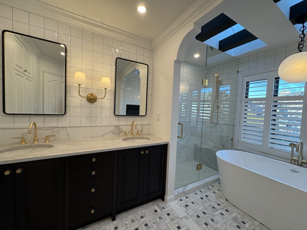 Modern bathroom with double black vanities, gold fixtures, two mirrors, a wall sconce, a glass shower, a freestanding tub, white tile walls, patterned tile floor, and large windows with white shutters.