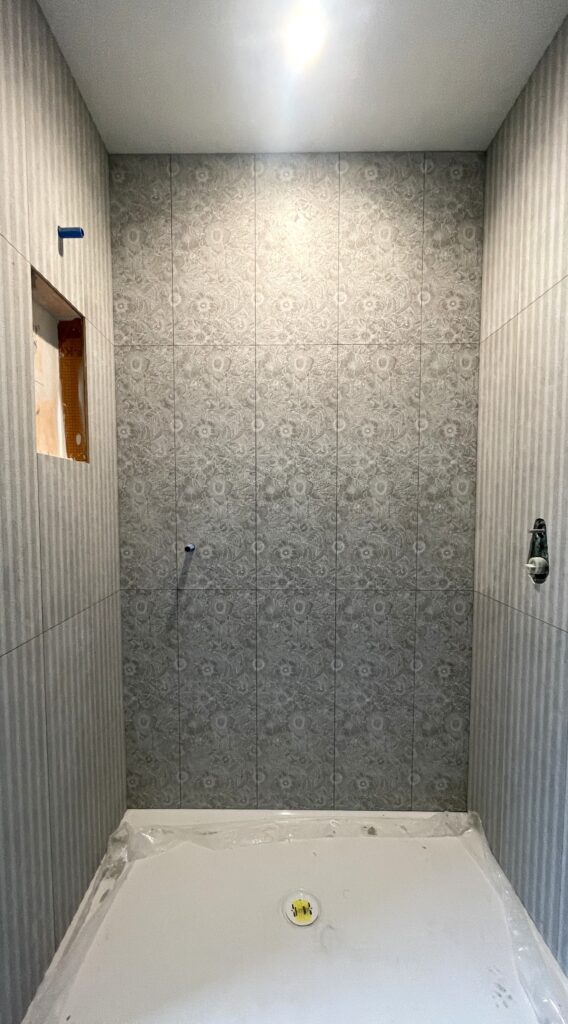 A shower under construction with patterned gray tiles on the back wall and striped gray tiles on the side walls. Fixtures and plumbing are exposed, and a protective sheet covers the shower floor.