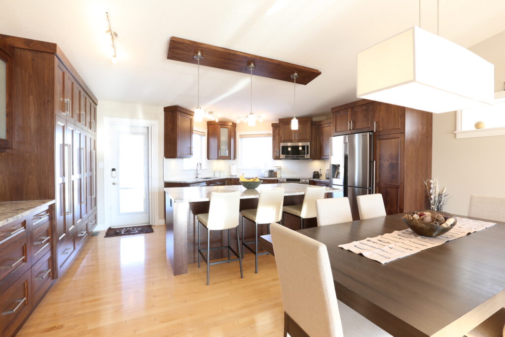 Modern kitchen and dining area with wooden cabinets, stainless steel appliances, a kitchen island with white barstools, and a dining table with beige chairs under a rectangular hanging light.