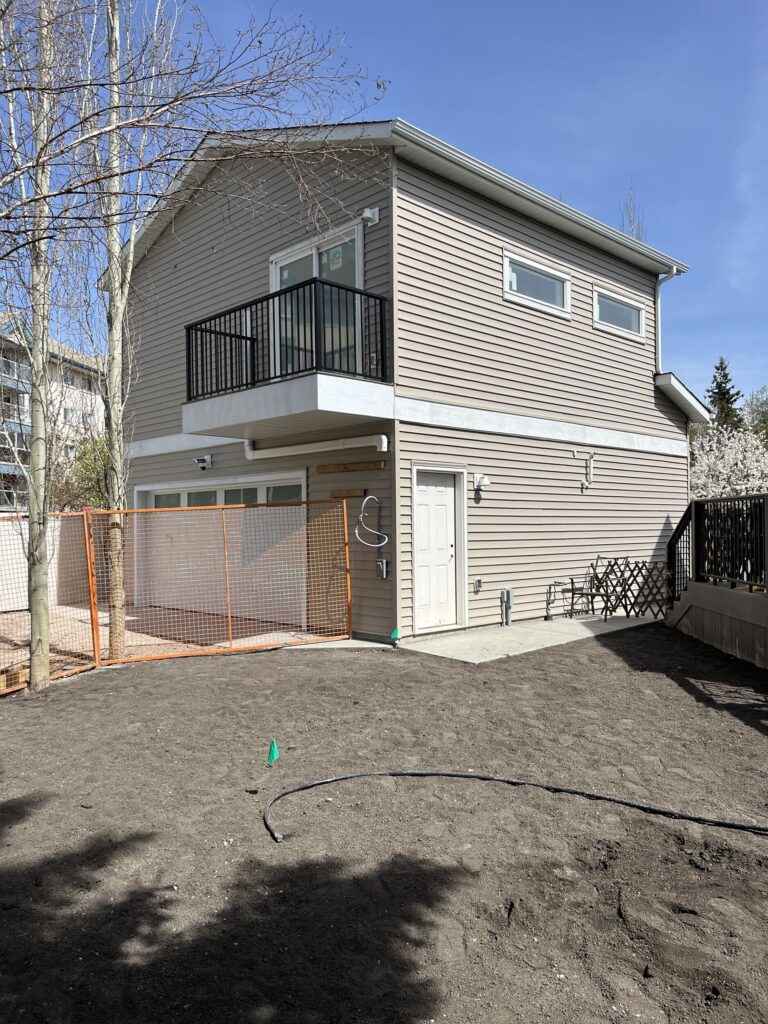 A two-story beige house with a balcony and white trim, next to a fenced area. The yard is bare soil with a garden hose, and leafless trees are nearby under a clear blue sky.