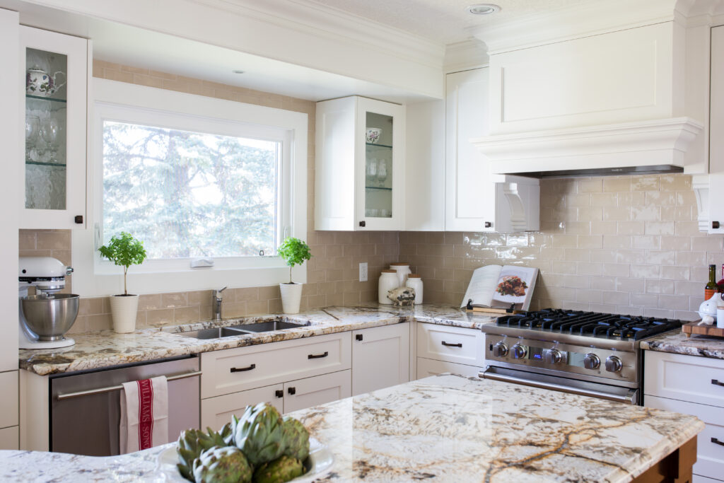 Bright, modern kitchen with white cabinets, granite countertops, a stainless steel stove, and a large window above the sink. Green plants and kitchen tools are neatly arranged, creating a clean and inviting atmosphere.