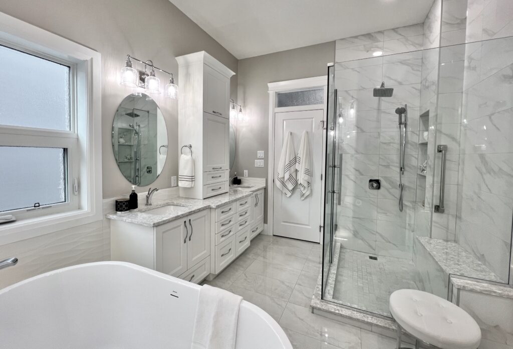 Modern bathroom with a freestanding bathtub, marble countertops, glass-enclosed shower, white cabinets, and gray walls. Towels hang by the door, and the room is brightly lit by wall-mounted lights and natural light.