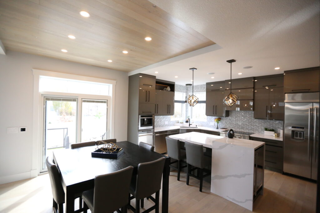 Modern kitchen with glossy gray cabinets, stainless steel appliances, a large island with white marble countertop, pendant lights, and a dining table with six chairs near glass doors letting in natural light.