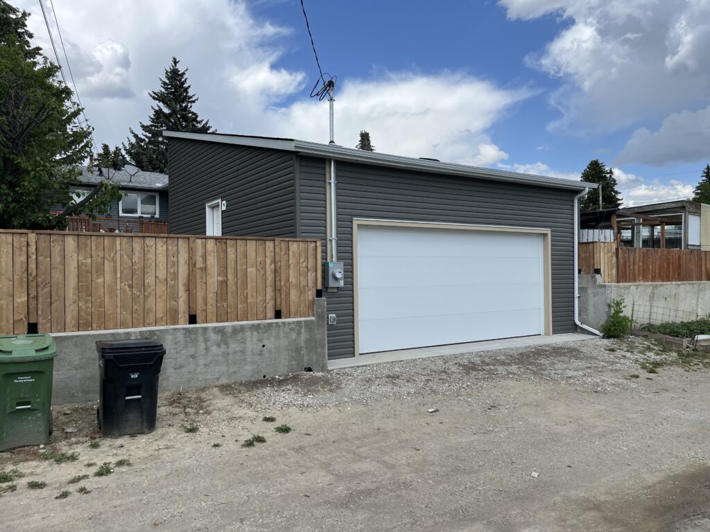 A two-car garage with dark gray siding and a white door sits next to a wooden fence. Trash and recycling bins are in front, and the sky above is partly cloudy. Gravel and dirt cover the alleyway beside the garage.