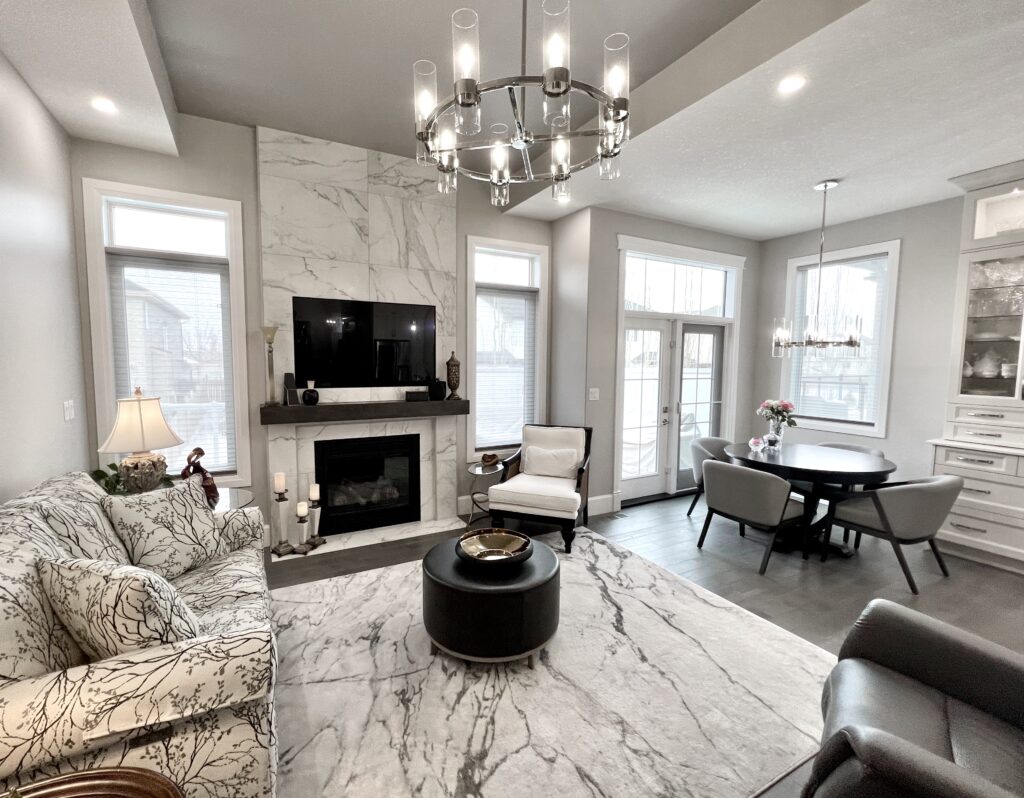 Modern living room with marble fireplace, wall-mounted TV, patterned sofa, armchair, round ottoman, chandelier, dining area with table and chairs, large windows, and neutral color scheme.