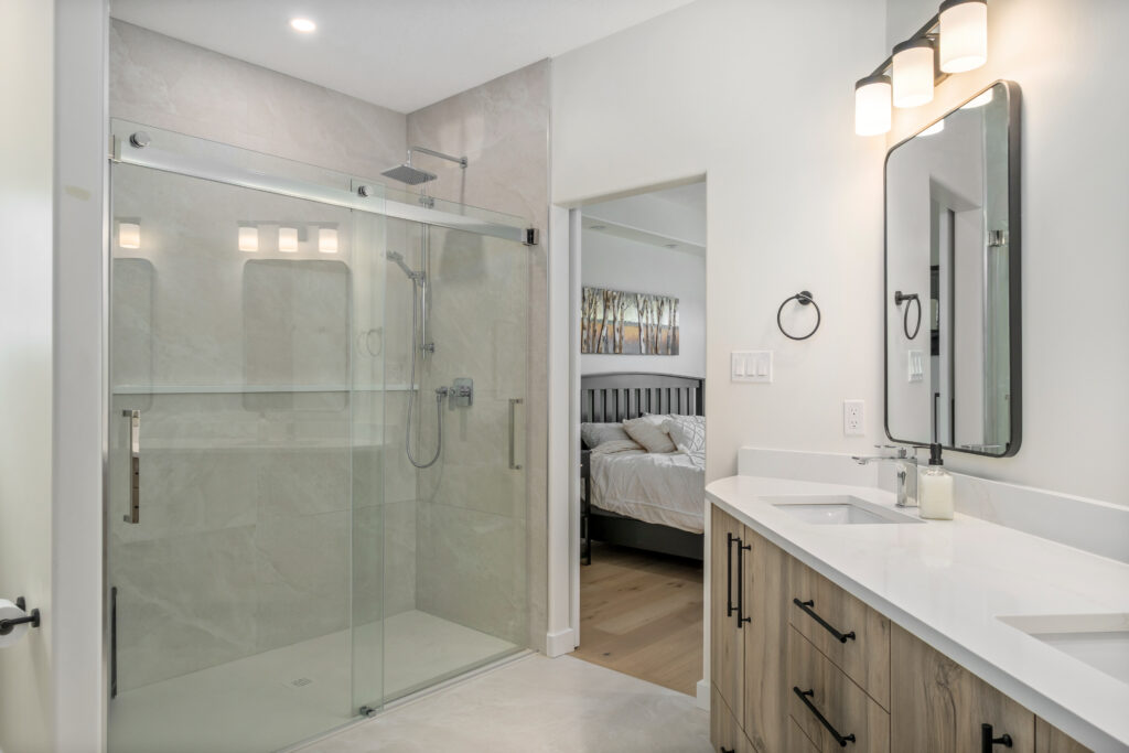 Modern bathroom with a glass-enclosed shower, white double-sink vanity with black hardware, wall-mounted lights, and a large mirror. An open doorway reveals a bedroom with a bed and wall art.