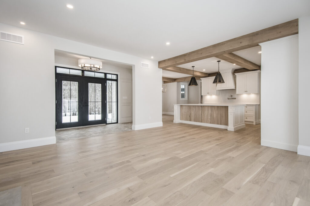 Spacious modern open-plan living area with light wood floors, white walls, exposed wooden ceiling beams, large black-framed glass doors, and a kitchen with white cabinets, island, and pendant lights.