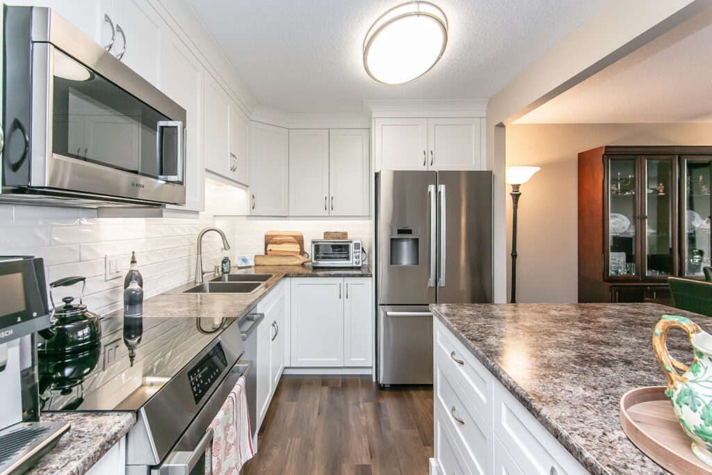 Modern kitchen with white cabinets, stainless steel appliances, marble-patterned countertops, and a dark wood floor. There are cutting boards and a toaster on the counter, with a lamp and dining area visible in the background.