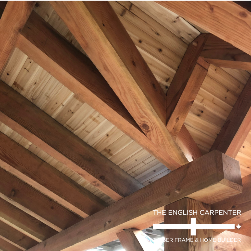 Wooden beams and planks form the underside of a pitched roof, showing detailed joinery. Sunlight highlights the wood grain. The English Carpenter logo and text appear in the lower right corner.