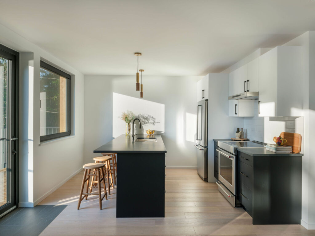 Modern kitchen with black and white cabinetry, a central island with wooden stools, stainless steel appliances, pendant lights, and large windows letting in natural light. A vase with flowers sits on the island.