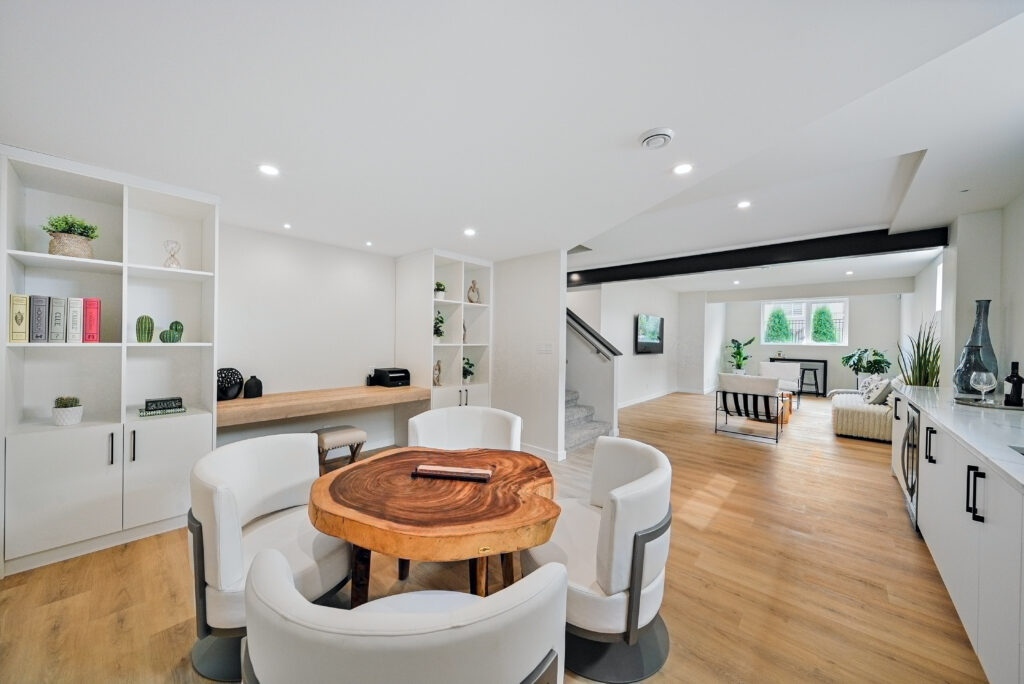 Modern open-plan living space with wooden floors, round wooden table with white chairs, built-in shelving, lounge area with sofas, and large windows letting in natural light.