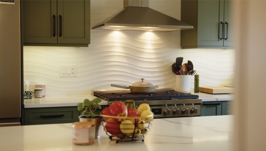 A modern kitchen with green cabinets, a stainless steel stove and hood, white wavy tile backsplash, and a fruit basket with apples, bananas, and lemons on a white countertop.
