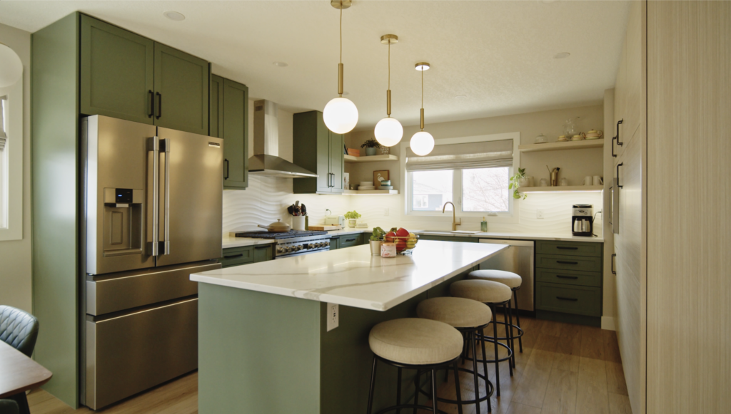 Modern kitchen with green cabinets, stainless steel appliances, white countertops, three round pendant lights, and three beige cushioned stools at an island. A window lets in natural light over the sink area.