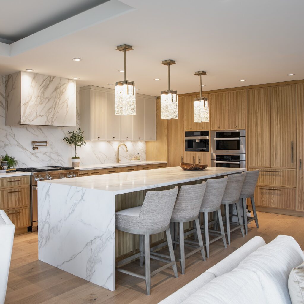 Modern kitchen with a large marble island, four gray upholstered barstools, wood cabinetry, built-in ovens, and three pendant lights hanging above the island. Light wood floors and neutral tones create a warm, elegant space.