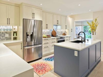 Modern kitchen with stainless steel fridge, white cabinets, patterned backsplash, and blue island with sink. Colorful floral rug and yellow flowers add bright accents. The space is well-lit and open.