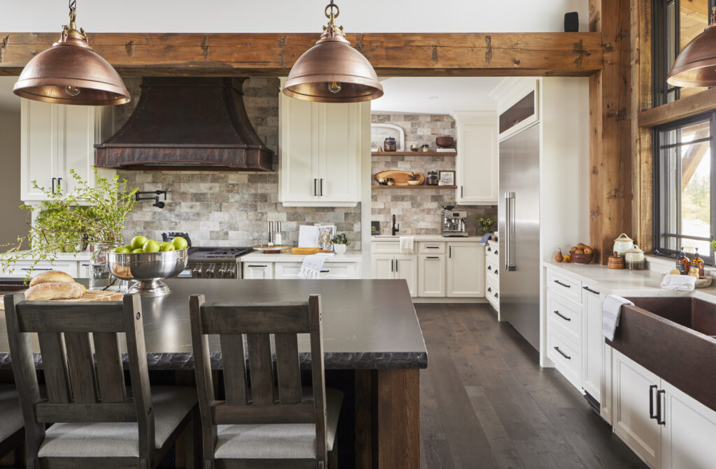 A rustic kitchen with wood beams, white cabinets, stainless steel appliances, a large island with dark countertop, wooden chairs, copper pendant lights, and a farmhouse sink by a window. Decorative items and greenery are on the counters.