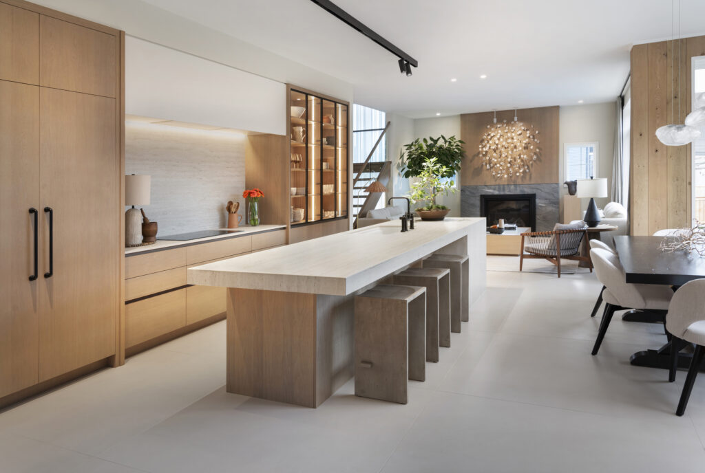 Modern open-concept kitchen and living area with light wood cabinetry, large stone island with four stools, built-in shelving, contemporary décor, and a cozy seating area near a fireplace.