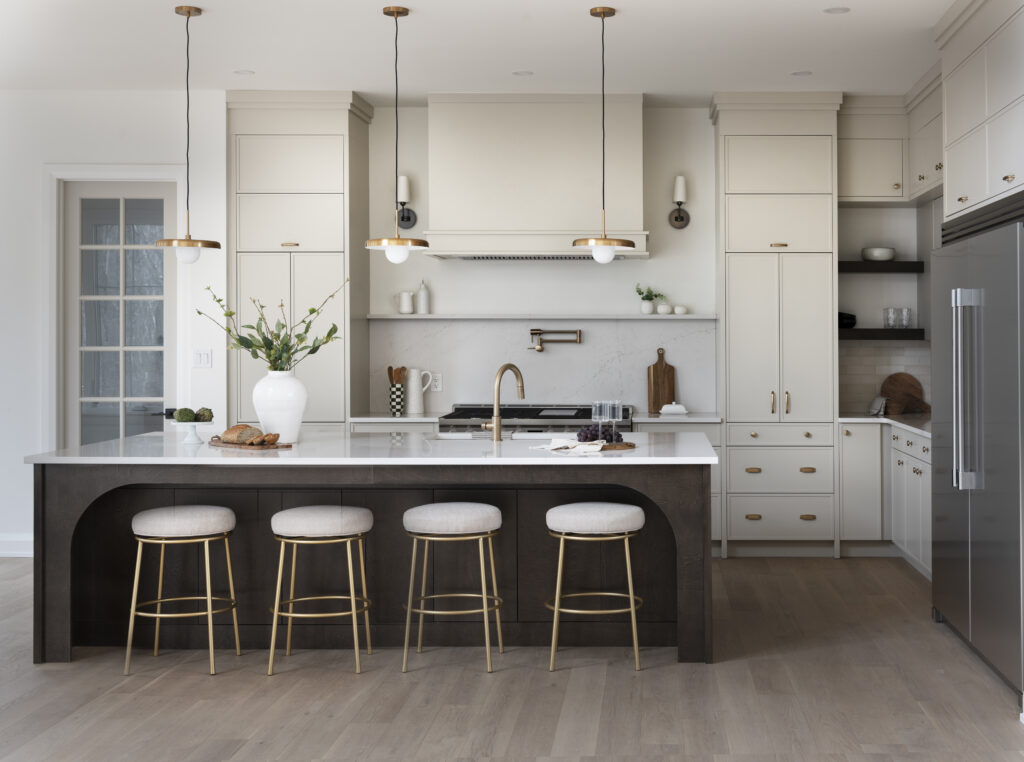 A modern kitchen with a large island featuring four round stools, pendant lights above, white cabinets, stainless steel appliances, and decorative accents like a white vase and cutting boards on the countertop.