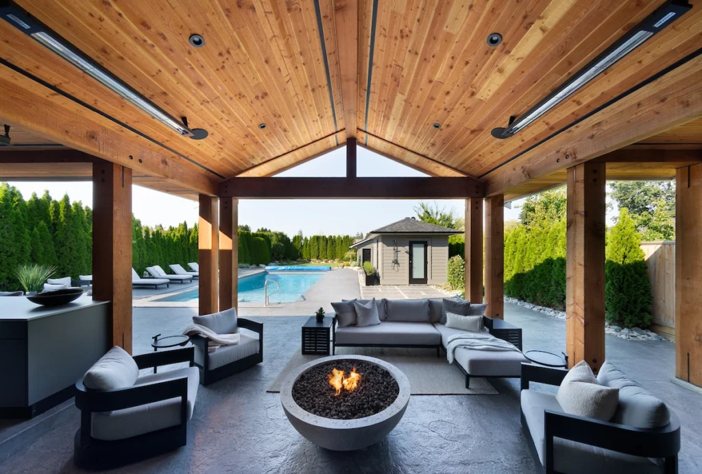 A covered outdoor patio with wooden beams features modern seating around a circular fire pit. Beyond the patio, there is a swimming pool surrounded by lounge chairs and a small pool house, enclosed by tall green hedges.