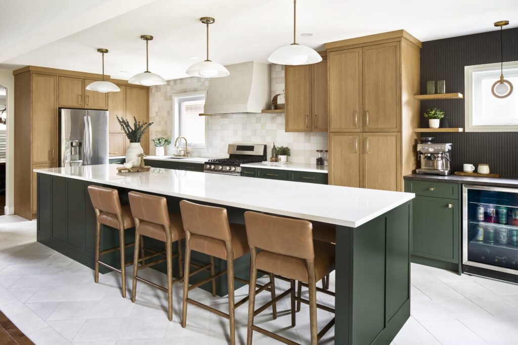 Modern kitchen with a large island featuring a white countertop, green cabinets, and four tan leather barstools. The space includes wood cabinetry, stainless steel appliances, and pendant lights above the island.