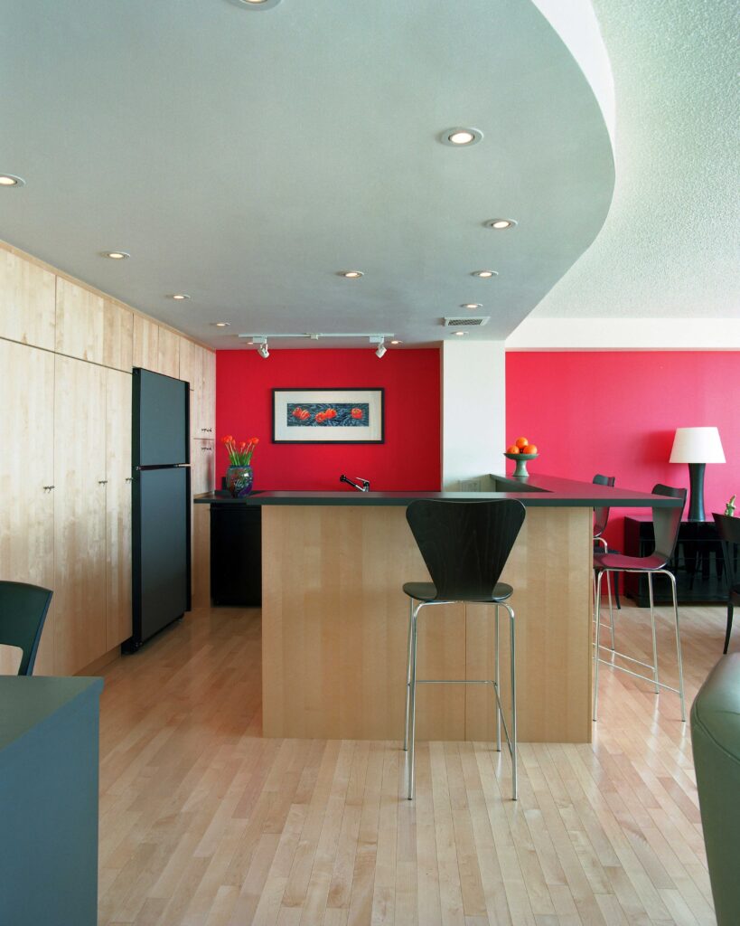 Modern kitchen with light wood floors and cabinets, a black fridge, and a center island with high chairs. Red accent wall with framed art and a vase of flowers on a side counter; track and recessed lighting overhead.