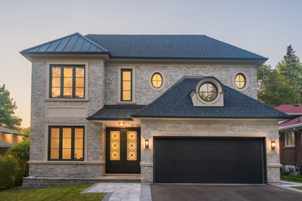 A modern two-story house with light brick exterior, large black-framed windows, a black double garage door, double front doors with glass panels, and exterior lights, photographed at sunset.