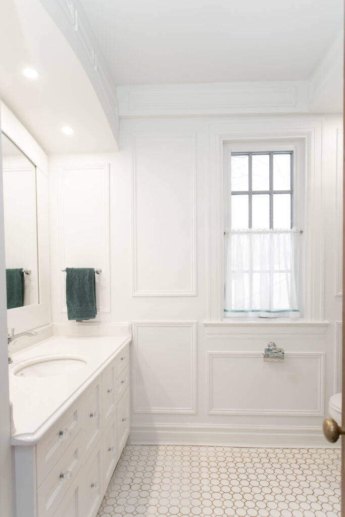 A bright, white bathroom with a large mirror above a sink and countertop, hexagon tile floor, a teal towel on a rack, and a window with white curtains letting in natural light.