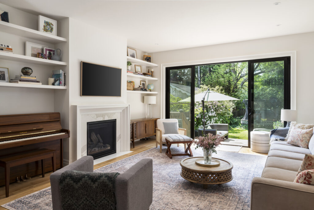 Bright living room with a fireplace, wall-mounted TV, shelves, piano, and comfy seating. Large glass doors open to a green patio with outdoor furniture and a garden umbrella. Flowers sit on a round coffee table.